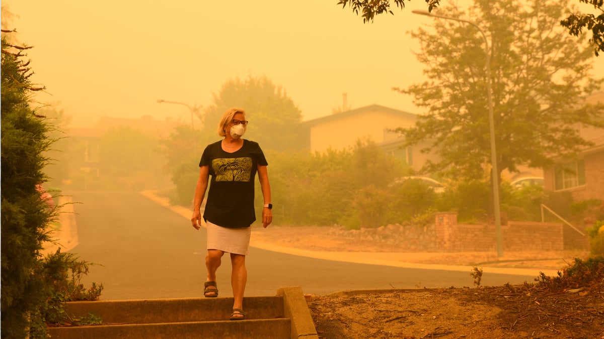 A woman wears a face mask with orange wildfire smoke seen around her. 